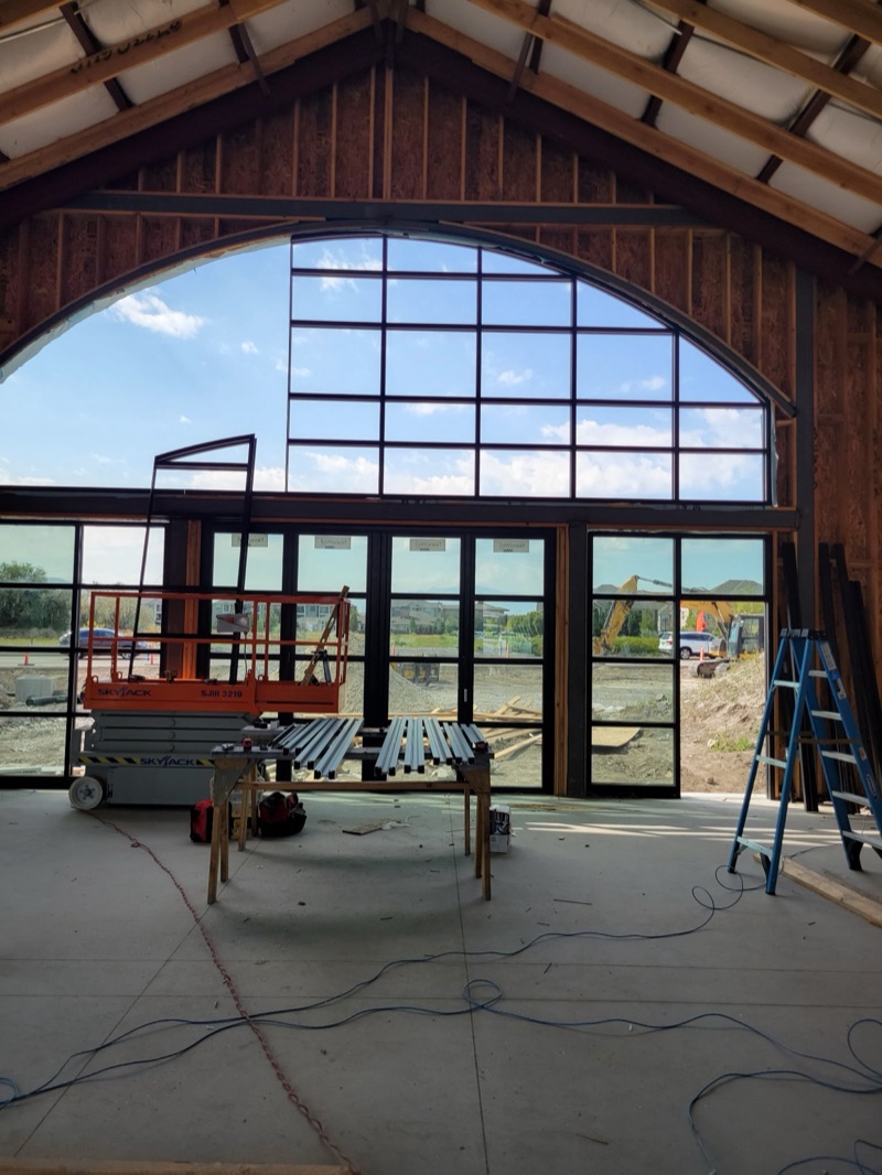A vaulted commercial interior looking out through a large arched window wall with dark aluminum framing, above a folding glass door system, with the building still in timber frame