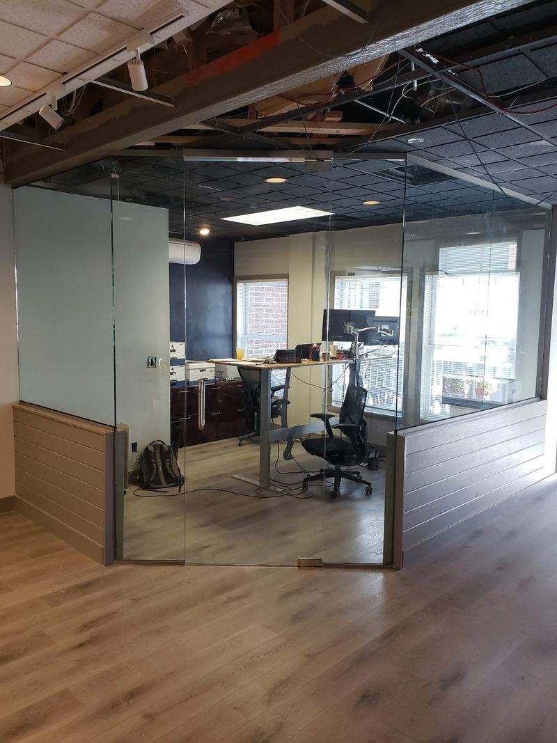A corner of a glass walled office with a frameless glass door and a low timber base
